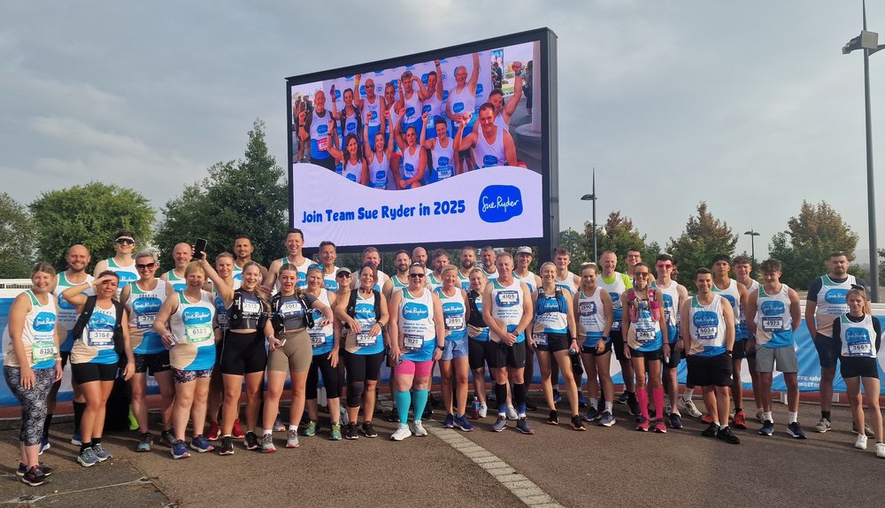 Lots of Sue Ryder runners in their race gear, beneath a video billboard showing celebrating runners