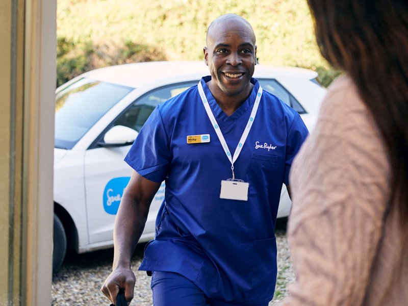 A Sue Ryder nurse in blue scrubs arrives at a patient's house. In the background is a Sue Ryder branded car and in the foreground is the back of the person opening the front door.