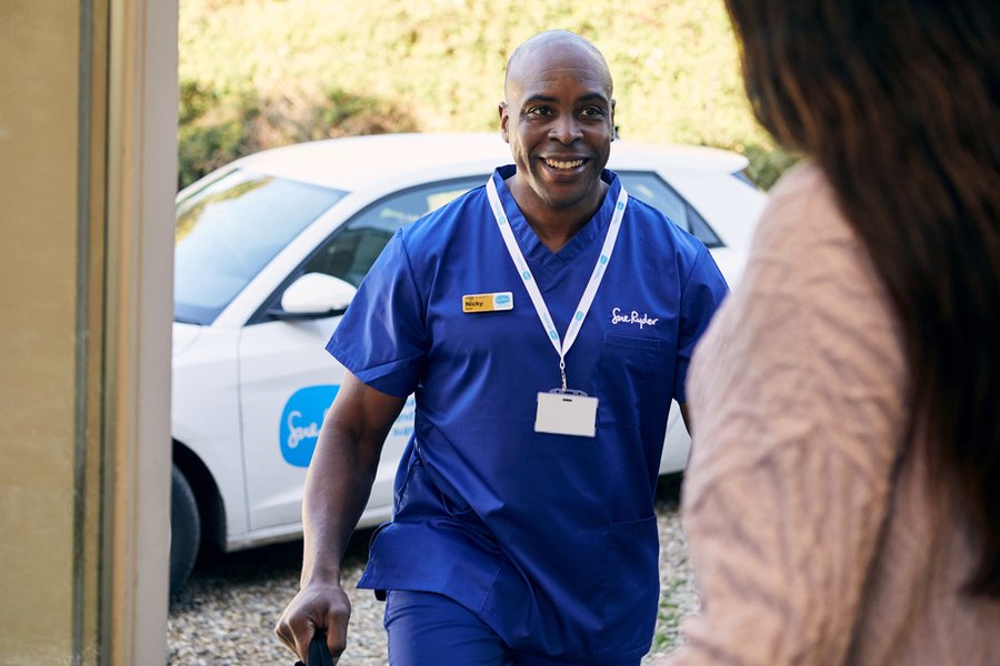 A Sue Ryder nurse in blue scrubs arrives at a patient's house. In the background is a Sue Ryder branded car and in the foreground is the back of the person opening the front door.