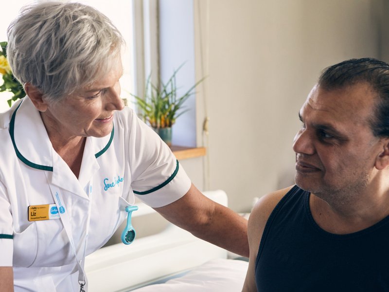 A nurse in a white uniform leans towards a patient sat on a bed, they are looking at each other and smiling slightly
