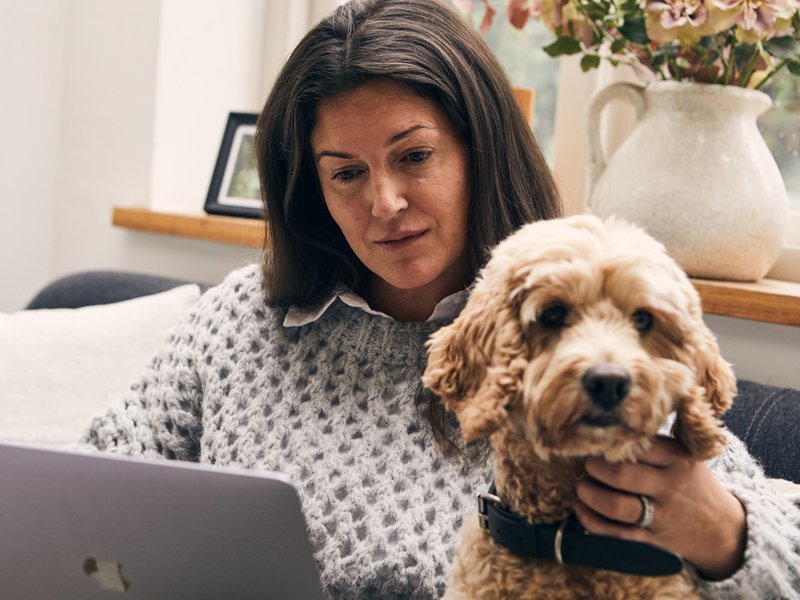 Woman sits on a sofa with an arm around her dog looking thoughtfully at her laptop. She is wearing a grey jumper and white shirt. In the background is a vase of flowers.