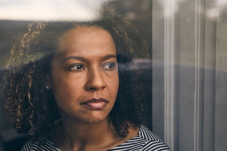 A woman with curly brown hair is staring out of a window with a thoughtful expression on her face