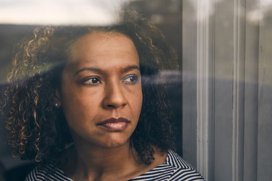 A woman with curly brown hair is staring out of a window with a thoughtful expression on her face