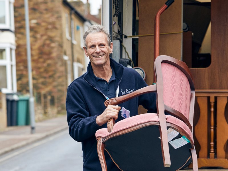 A delivery driver stands in front of a Sue Ryder van holding a chair he has collected from a donor