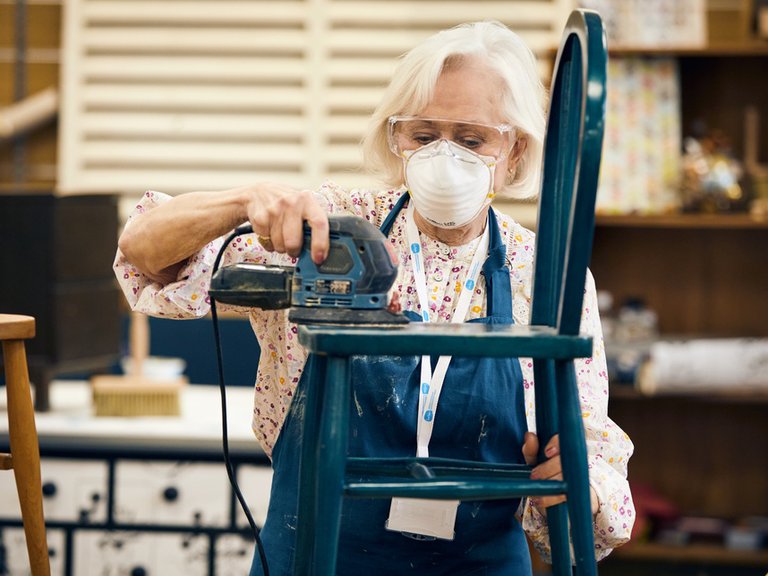 A woman with a protective face mask is sanding down a blue wooden chair.