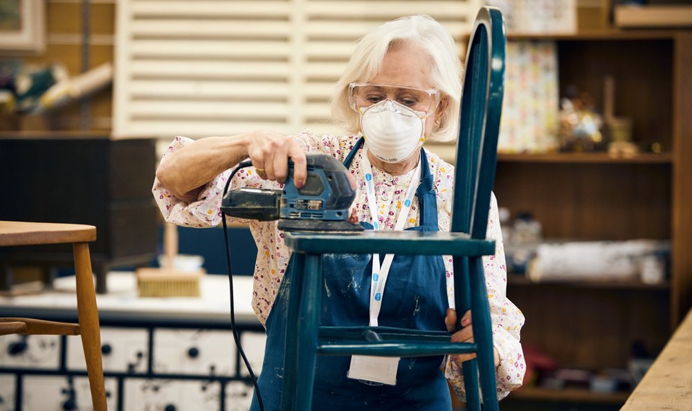 A woman with a protective face mask is sanding down a blue wooden chair.