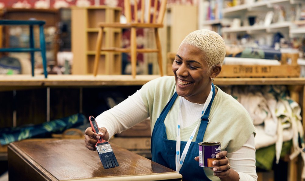 A woman in a blue apron is varnishing the top of a table in a Sue Ryder workshop.