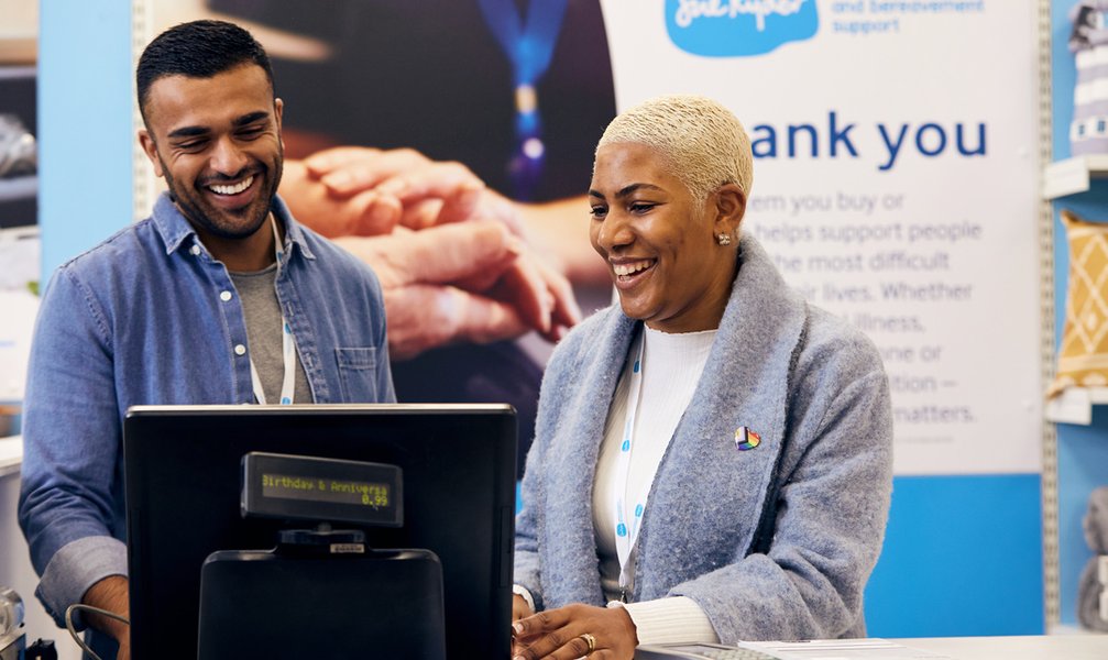 A male and female volunteer stand behind the till in a Sue Ryder charity shop