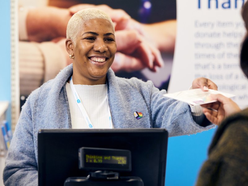 A smiling shop staff member is stood behind the till and handing a receipt to a customer, who's back you can see.