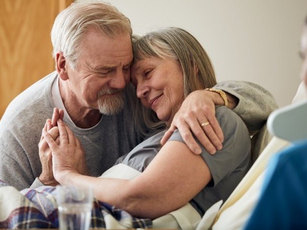 A patient sits up in bed, hugging her family member to the side. They are both smiling. In the foreground is the side profile of a Sue Ryder Nurse.