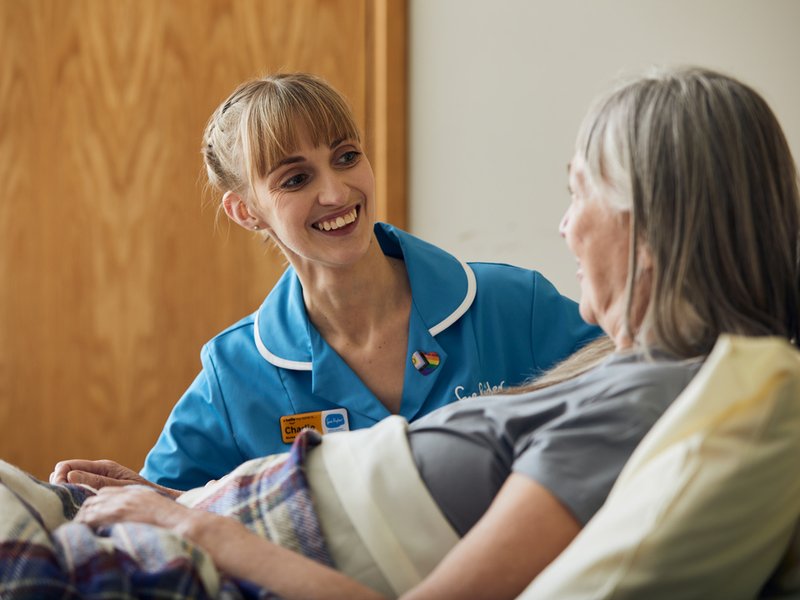 A nurse in a light blue Sue Ryder uniform smiles at a female patient sat up in a bed.
