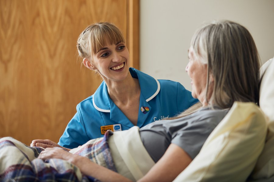 A nurse in a light blue Sue Ryder uniform smiles at a female patient sat up in a bed.