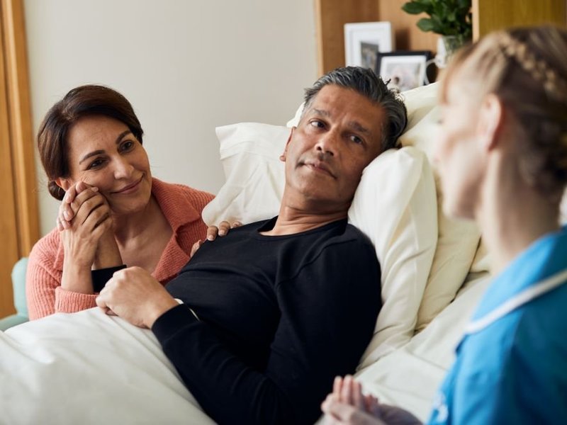 A patient is sat up in bed, holding hands with a family member sat next to them. In the foreground is a nurse, talking to them.