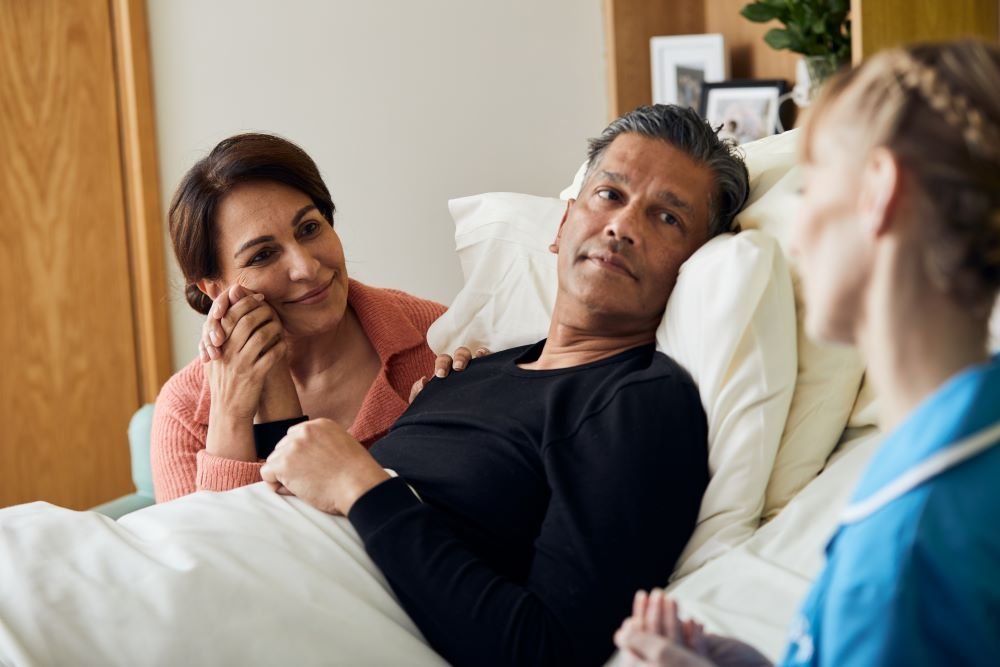 A patient is sat up in bed, holding hands with a family member sat next to them. In the foreground is a nurse, talking to them.