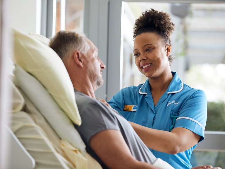 A female nurse in a light blue uniform stands next to the bed of a male patient. The bed is raised so both people are at the same height and are looking at each other.