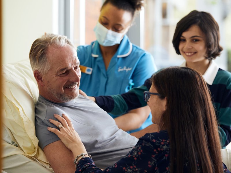 A woman and child are next to a patient's bedside, they are all looking at each other and smiling. A nurse is in the background with a face mask on.