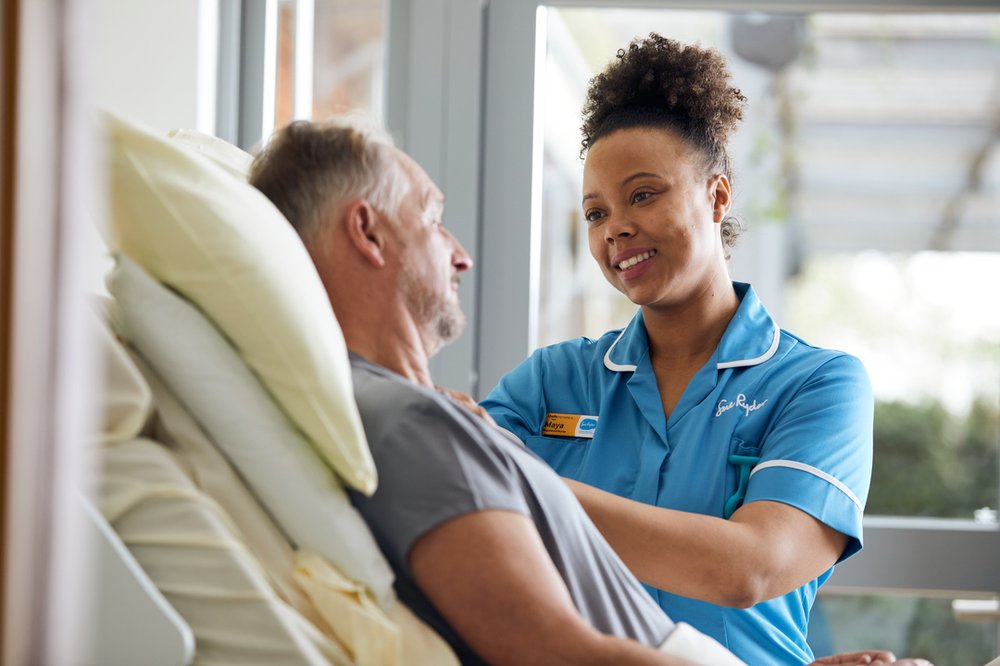A female nurse in a light blue uniform stands next to the bed of a male patient. The bed is raised so both people are at the same height and are looking at each other.