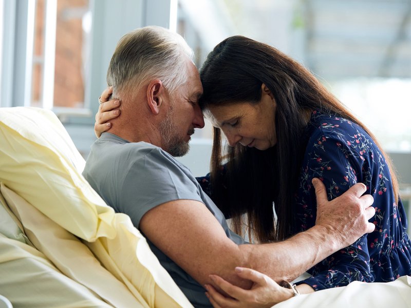 A woman sits on the edge of a hospice bed and embraces the male patient in a comforting gesture. Their heads are together.