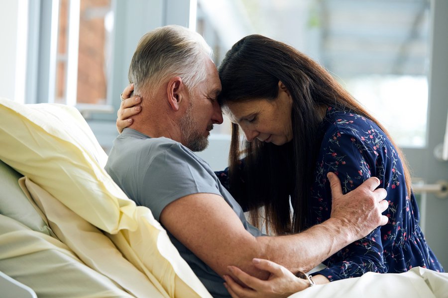 A woman sits on the edge of a hospice bed and embraces the male patient in a comforting gesture. Their heads are together.