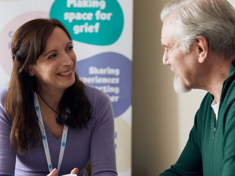 A man with grey hair looks at a woman with dark hair wearing a Sue Ryder lanyard. In the background is a poster advertising our Grief Kind Spaces.