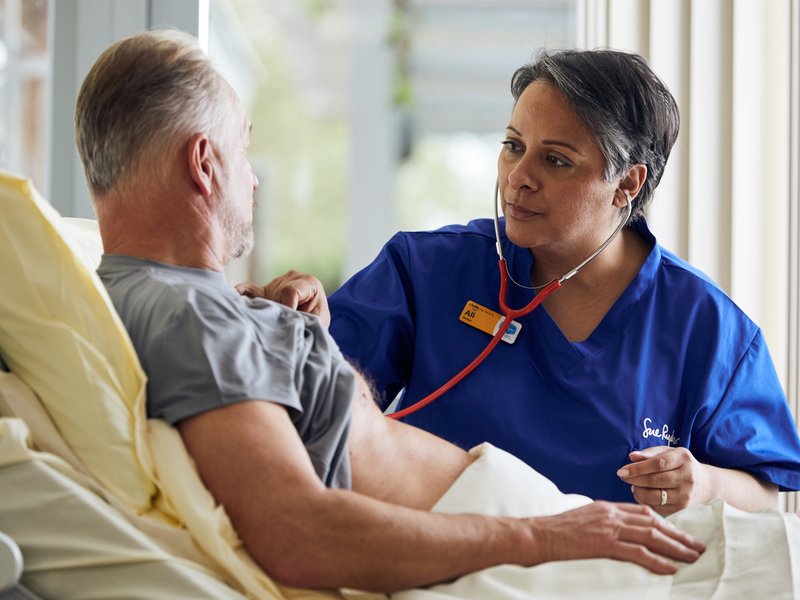 A doctor in blue scrubs is stood next to a patient's bed, using a stethoscope to listen to the patient's heart.