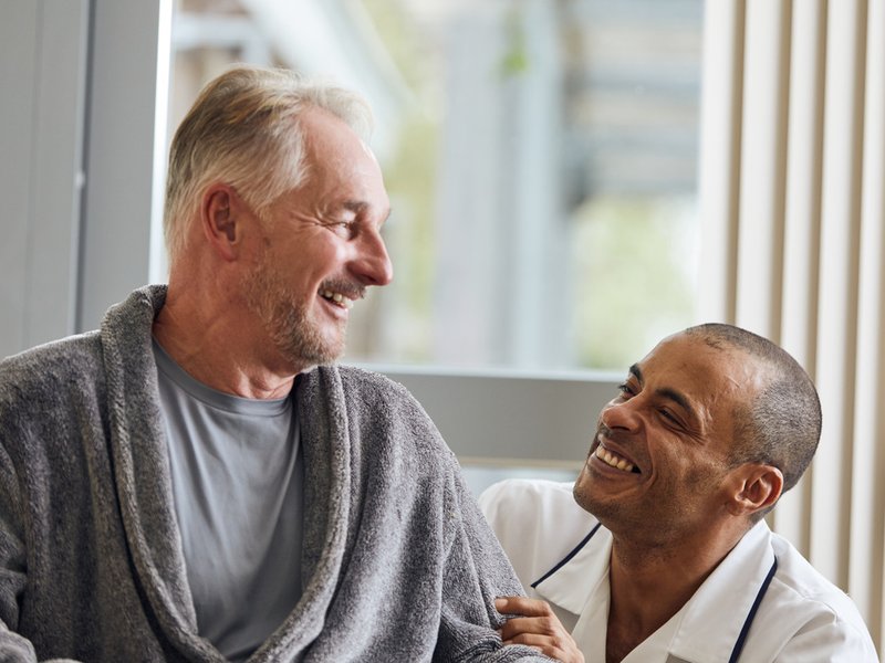A patient in a grey dressing gown is sat down with a physio in a white uniform crouched beside him. They are smiling at each other.