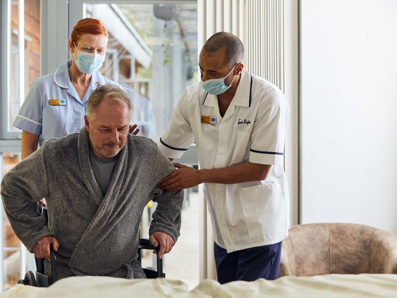 A patient is getting out of a wheelchair with the help of a physio. A nurse is in the background holding the wheelchair.