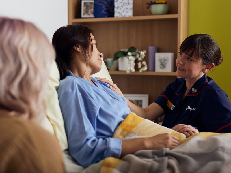 A nurse in a dark blue uniform is sat next to a bed, where a female patient is sitting. In the foreground is the back of another woman.