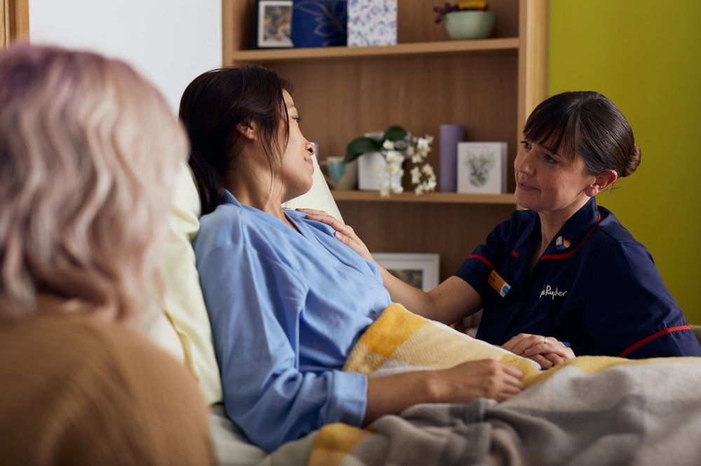A female Sue Ryder Nurse sitting next to a female patient who is in bed. The Nurse has a hand on the patient's shoulder and is talking to her. A female relative is also present at the patient's bedside.