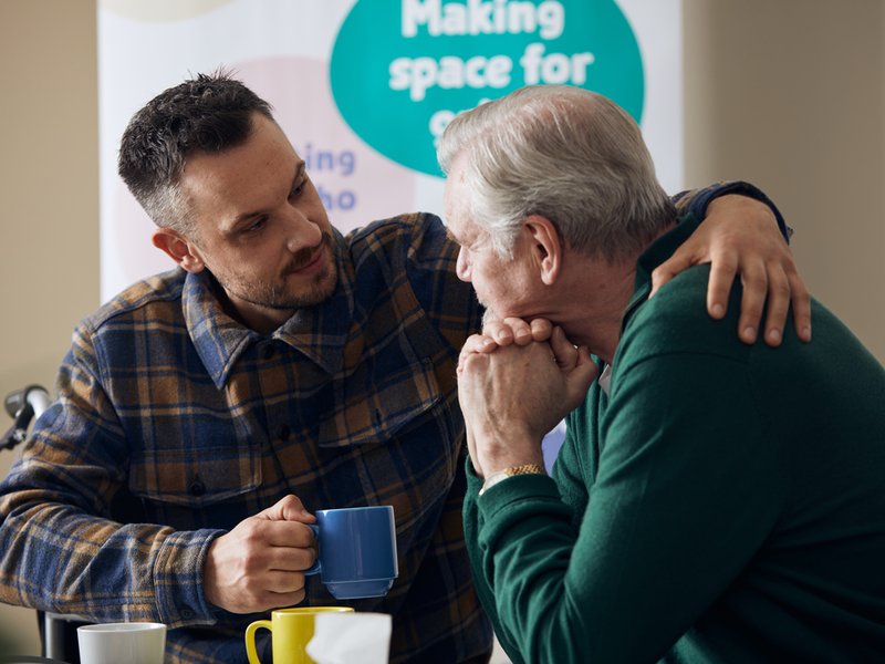 Two men are sat with mugs, the younger man has his arm around the older man in a comforting gesture