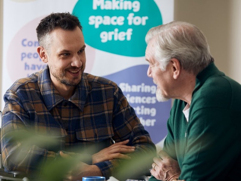 Two men, one older, are looking at each other and chatting. In the background is a Sue Ryder Grief Kind Space poster