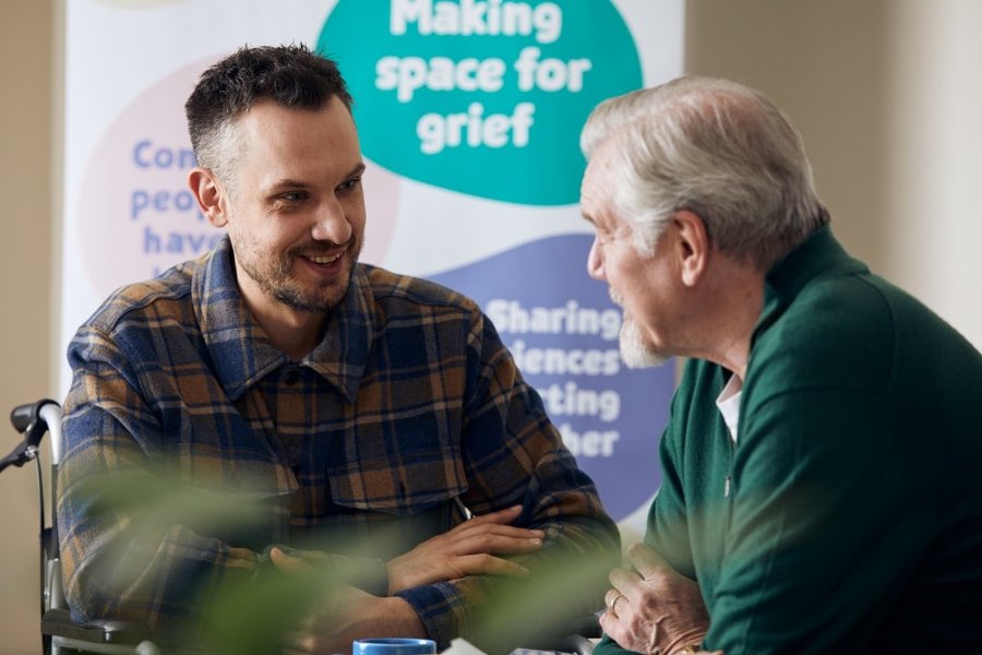 Two men, one older, are looking at each other and chatting. In the background is a Sue Ryder Grief Kind Space poster