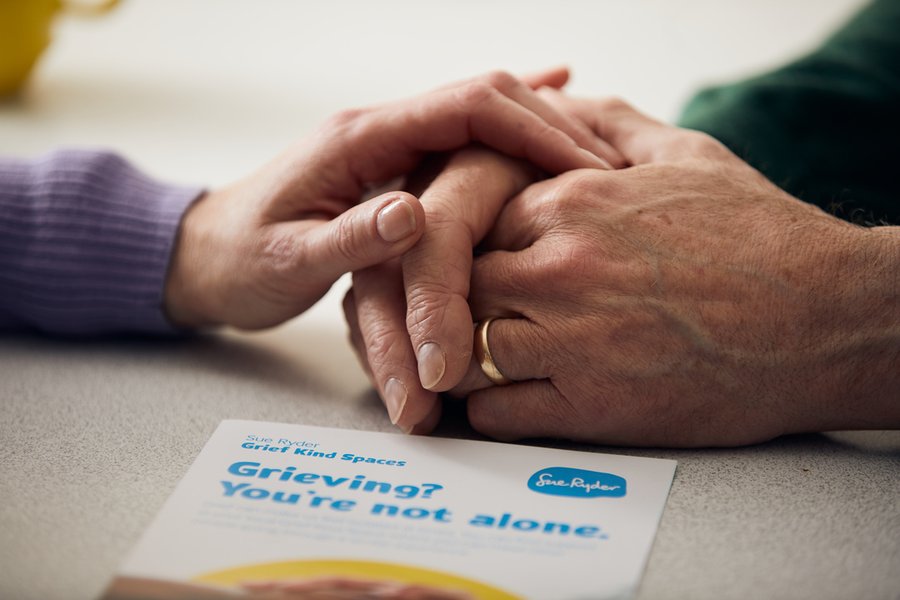 A person rests their hand on another's clasped hands. Next to the hands is a leaflet about grief.