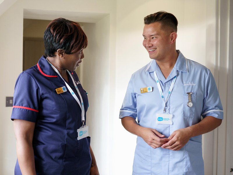 One female Sue Ryder Clinical Nurse Specialist and one male Healthcare assistant chatting and laughing with one another in a Sue Ryder hospice