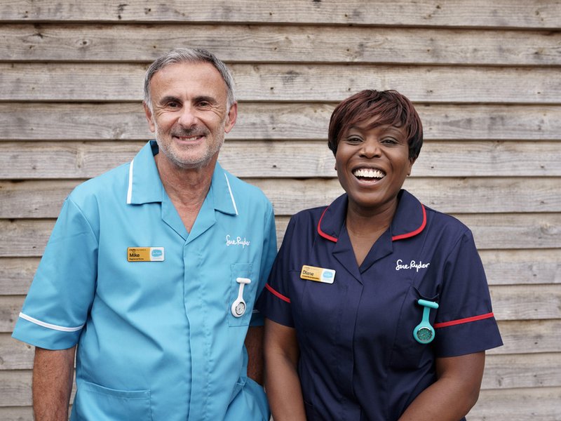 One female Sue Ryder Clinical Nurse Specialist and one male Sue Ryder Nurse smile at the camera, standing next to one another