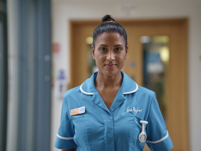 A nurse in a blue Sue Ryder uniform smiles directly at the camera
