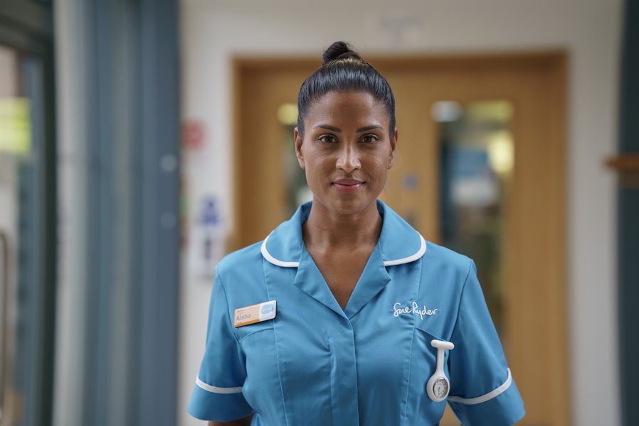 A nurse in a blue Sue Ryder uniform smiles directly at the camera