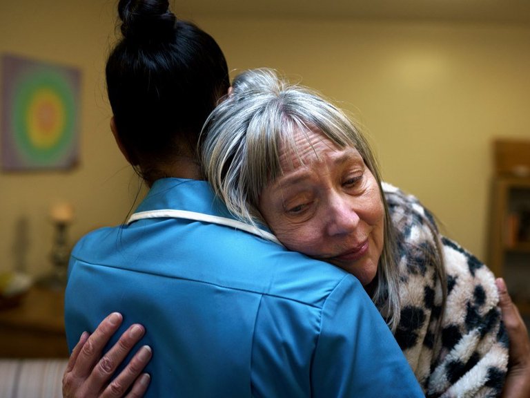 A tearful woman lays her head on the shoulder of a Sue Ryder Nurse  who embraces her.