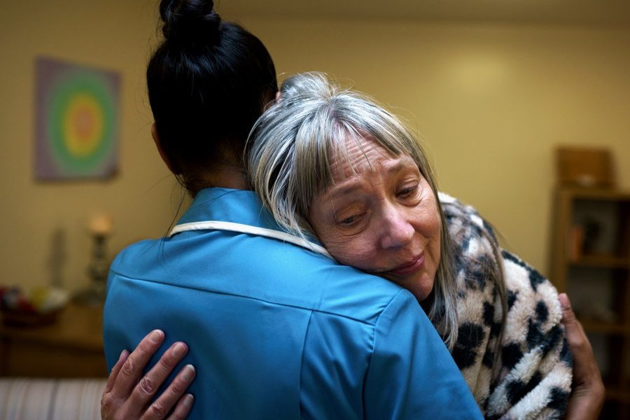 A tearful woman lays her head on the shoulder of a Sue Ryder Nurse  who embraces her.