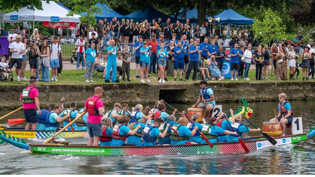 Teams in dragon boats, racing along a river, with crowds cheering behind them
