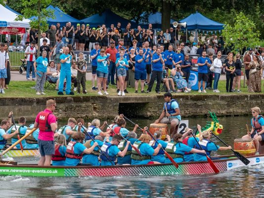 Teams in dragon boats, racing along a river, with crowds cheering behind them