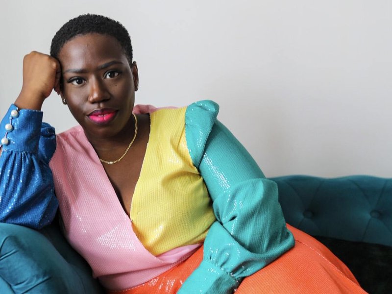 Candice is sat on a teal sofa looking directly at the camera. She is wearing a multi-coloured dress.