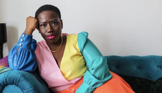 Candice is sat on a teal sofa looking directly at the camera. She is wearing a multi-coloured dress.