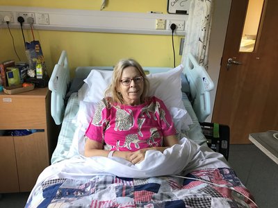 Female patient sits up in her bed in a Sue Ryder hospice. She is smiling at the camera and wearing a bright pink t-shirt with cheetahs on it.