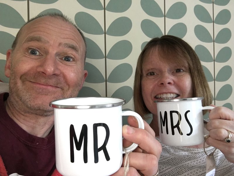 David and Sarah are smiling at the camera and holding up mugs with 'Mr' and 'Mrs' written on them.