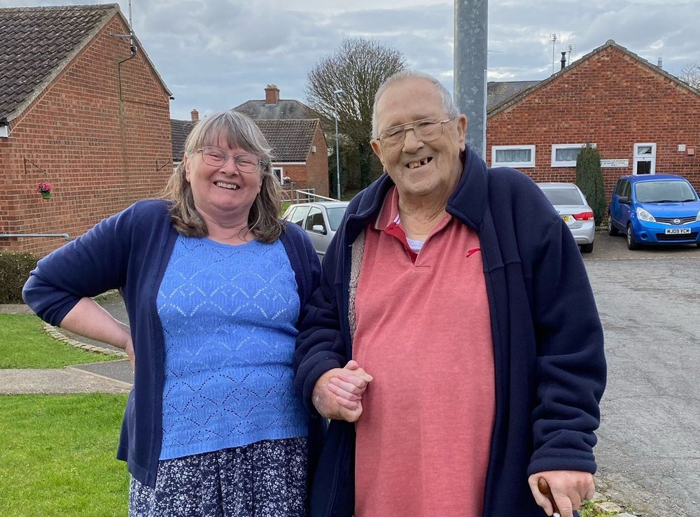 Debbie and Doug hold hands as they smile into the camera. In the background there are a few cars and some bungalows.