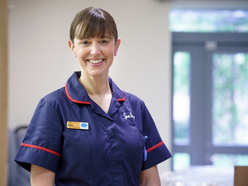 A senior nurse in a dark blue uniform is smiling at the camera.