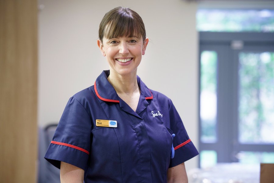 A senior nurse in a dark blue uniform is smiling at the camera.