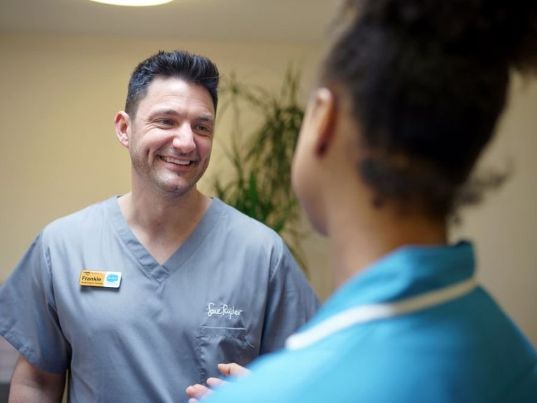 A healthcare worker in grey scrubs is talking to a nurse in a blue uniform.