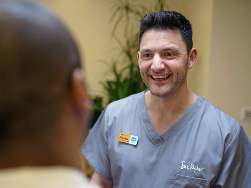 A healthcare volunteer in grey scrubs is smiling at another team member. We can see the back of their head in the foreground.